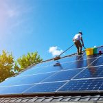 Person cleaning solar panels on a residential rooftop using a soft brush, surrounded by cleaning supplies and a ladder, representing regular maintenance for solar energy efficiency.